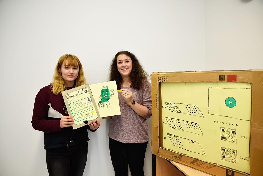 Nicole Vasiljuk (links) und Lisa Akili studieren beide Theater- und Medienwissenschaften. Gemeinsam mit Lukas Kuhn arbeiten sie an der Idee einer Handy-App, die Studierenden helfen soll, mit Hilfe von QR-Codes in Bibliotheken leichter einen Lernplatz zu finden. Zur besseren Visualisierung der einzelnen Menüpunkte haben sie aus Karton und Papier ein I-Pad sowie einen Bildschirm gebastelt. (Bild: FAU/Rebecca Kleine Möllhoff)
