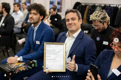 Zwei Männer in Bildmittelpunkt in Menge aus Menschen auf Stühlen, einer hält eine EELISA Urkunde und zeigt den Daumen hoch