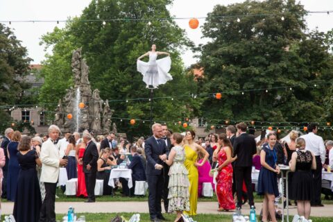 Schwebende Performerin auf dem Schlossgartenfest 2024 der Friedrich-Alexander-Universität Erlangen-Nürnberg. Zu sehen ist eine schwebende Artistin in weißem lockerem Kostüm im Hintergrund. Im Vordergrund Gäste des Fests, Girlanden und der Brunnen des Schlossgartens.