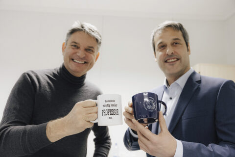 Rosshart und Hornegger halten Tassen hoch. eine tasse mit FAU -logo, die ander mit dem Aufdruck: So sieht ein richtig cooler Professor aus.