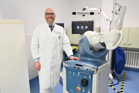 Prof. Marcel betsch, ein mann mittleren alters mit Platte und grauem Haarkranz sowie Brille, steht stolz grinsend bei dem roboter.