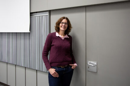 Frau Arntz, eine Frau mittleren Alters mit rotbraunen Haaren, steht an der Wand angelehnt, die Hände in den Hosentaschen.