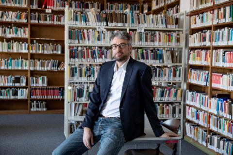 Kirchner, ein Mann mittleren Alters mit grauen Strähnen im Haar und einer Brille, sitzt auf einem Tische in einer Bibliothek.