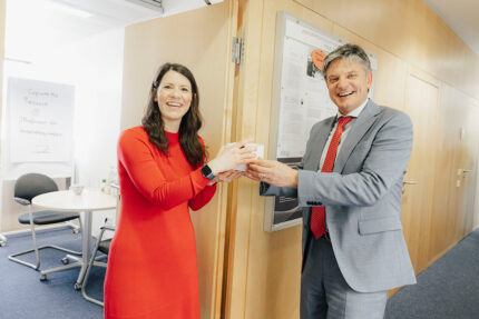 Hornegger und Handke, eine junge Frau mit dunklen Haaren und in rotem Kleid, halten gemeinsam ein Gastgeschenk von Hornegger.