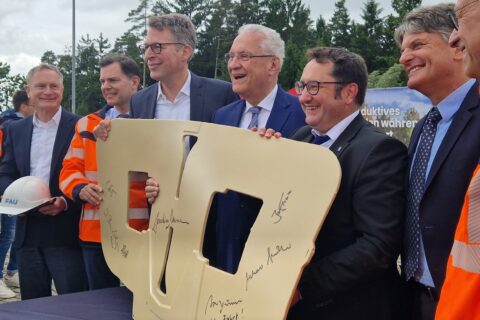 Sechs Männer in Anzügen halten einen beigen Nachbau einer Induktionsspule mit Unterschriften darauf