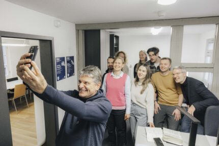 Präsident Hornegger und das Team von Prof. Necker am ifo stehen gemeinsam in einem Raum. Hornegger steht im Vordergrund und nimmt, gemeinsam mit den Leuten hinter ihm, ein Selfie auf.
