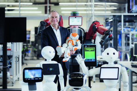 Franke und einige roboter aus dem Faps. Er grinst und trägt einen kleinen Roboter wie ein kind auf dem Arm.