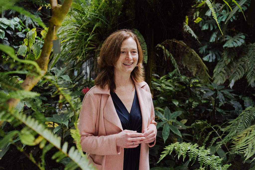 Prof. Jansen, eine Frau mit braunen haaren, trägt eine pinke lederjacke und steht im Botanischen Garten vor Bäumen und sträuchern.