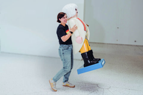 Laura Kneppe, eine Frau mit kurzen dunklen Haaren und Brille, trägt eine übergroße Playmobilfigur.