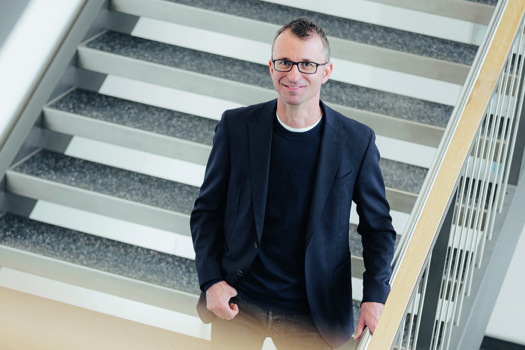 Dr. Wieland, ein Mann mit Brille, geheimratsecken und im schwarzen Jacket, lächelt aus der Vogelperspektive in die Kamera.