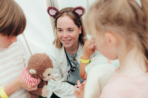 Frau Bittener, eine junge frau mit braunen Haaren, trägt eine Arztkittel und einen Haarreif mit Bärenohren. sie erklärt Kinder etwas.