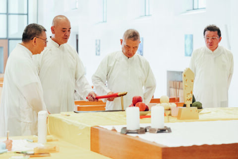 Drei asiatische Männer in weißen Roben mit einem Gerät aus rotem Holz an einem Altar.