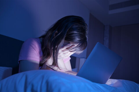 Frau hält sich die Händer vors Gesicht, sie sitzt im Dunkeln auf dem Bett, ihr Bild wird vom Laptopbildschirm vor ihr erleuchtet